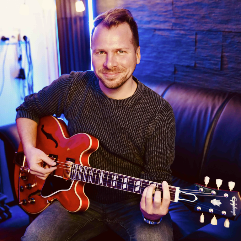 Musikproduktion Coaching in Hamburg Mann spielt eine rote Gitarre in einem gemütlichen Raum mit sanfter Beleuchtung.